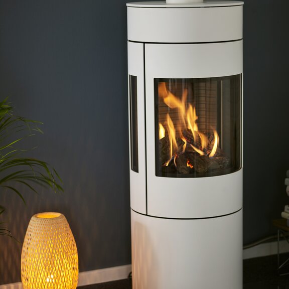 Stufa a gas VIVA 120 L in bianco con porta in acciaio e finestre laterali davanti a una parete blu scuro accanto a una lampada da terra accogliente