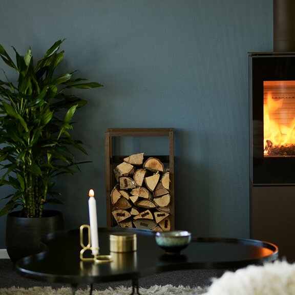 Stufa a legna NEXO 120 in nero con porta del focolare in vetro e fuoco silenzioso davanti a una parete color petrolio in una casa di stile